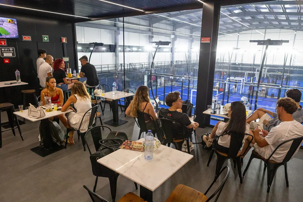 O bar cheio, com mesas brancas junto à parede de vidro e os campos azuis lá em baixo.